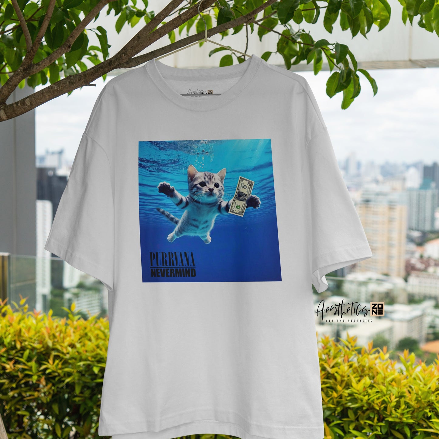 White t-shirt with a graphic of a cat in water and text, hanging outdoors with greenery and a cityscape in the background.
