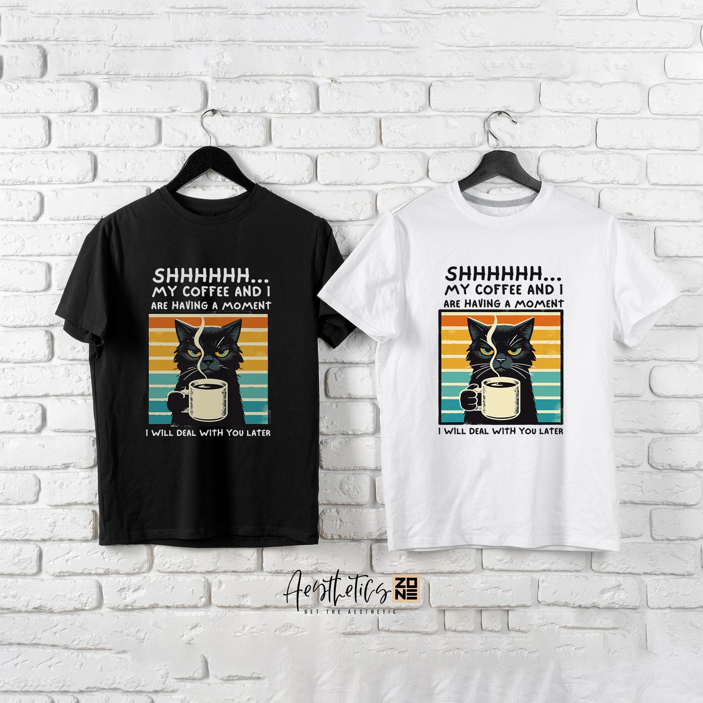 Black and white t-shirts with cat and coffee design on hangers against a white brick wall.