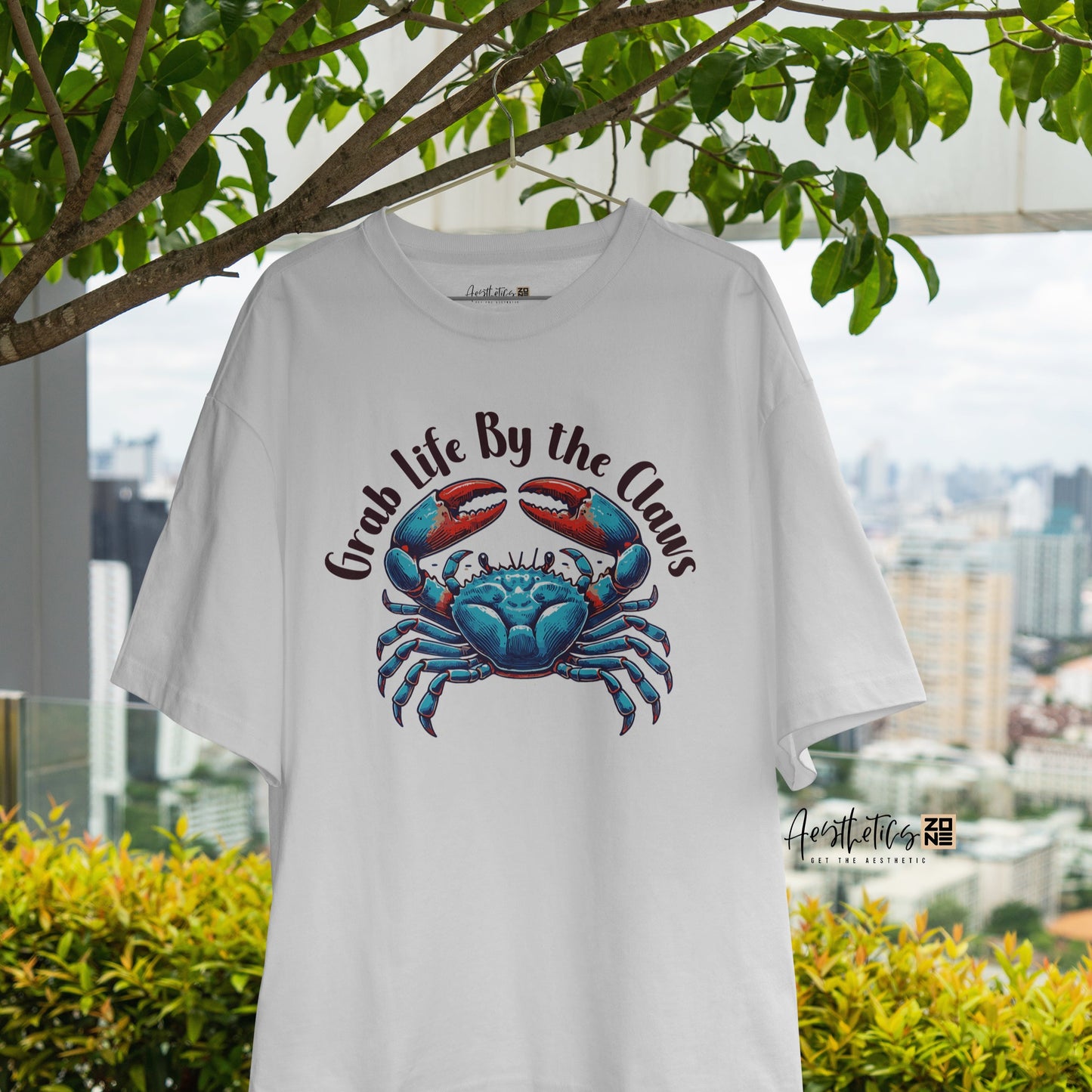 White t-shirt with a blue crab graphic and text, hanging outdoors with greenery and cityscape in the background.