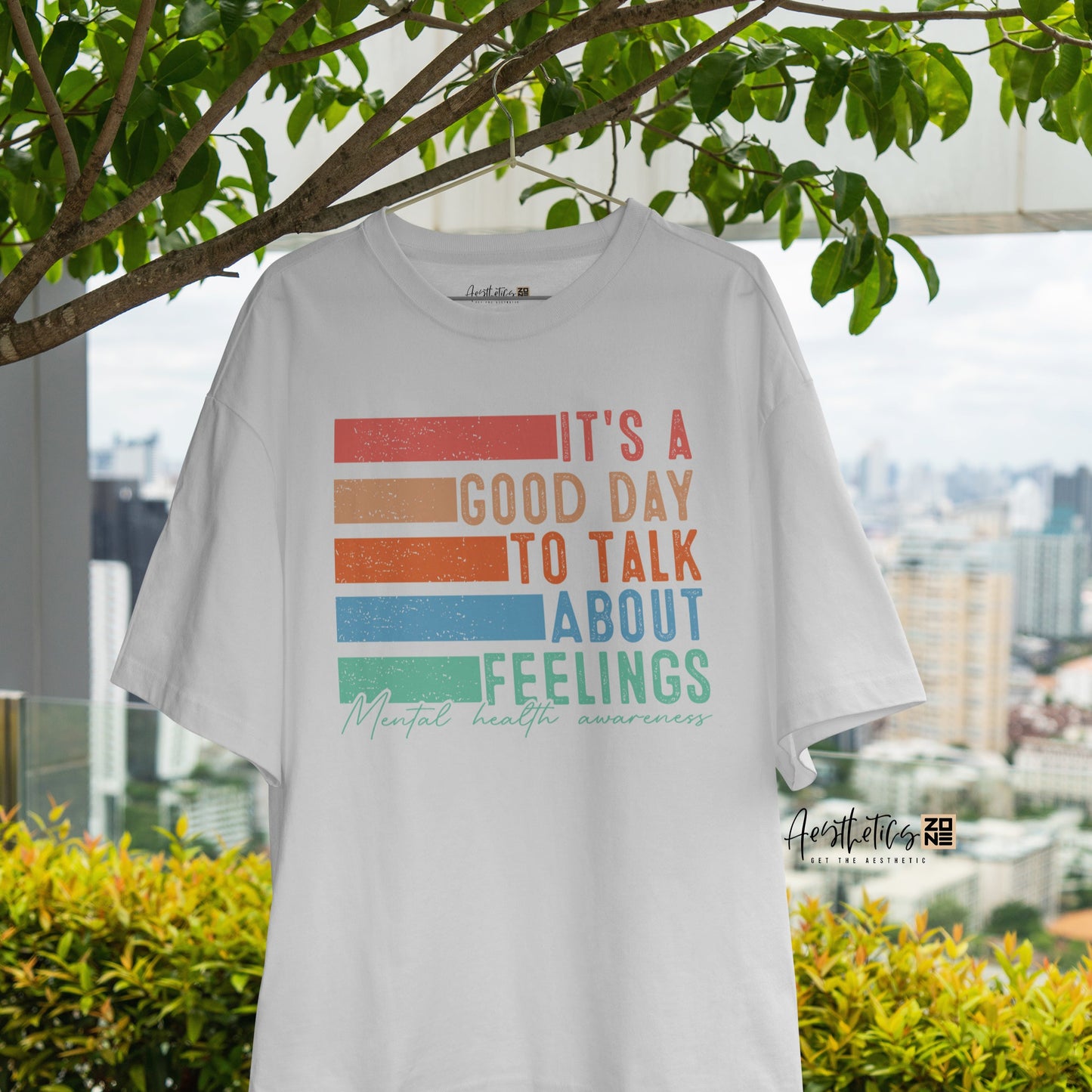 White t-shirt with colorful text design hanging outdoors with greenery and a cityscape in the background
