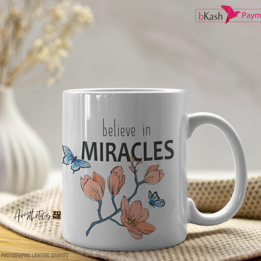 White mug with 'believe in miracles' text and floral design on a table.