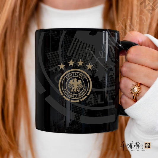Black mug with a gold emblem held by a person with red hair.