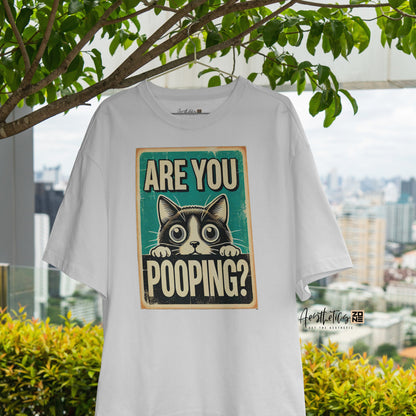 White t-shirt with a graphic of a cat and text 'Are you pooping?' hanging outdoors with greenery and a cityscape in the background.