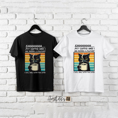 Black and white t-shirts with cat and coffee design on hangers against a white brick wall.