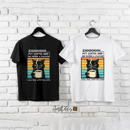 Black and white t-shirts with cat and coffee design on hangers against a white brick wall.