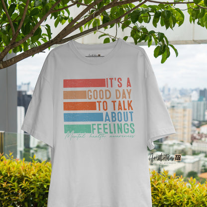 White t-shirt with colorful text design hanging outdoors with greenery and a cityscape in the background